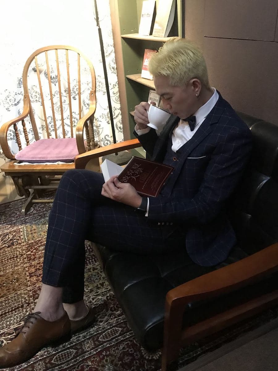 坐在店內沙發上的男士,穿深藍格紋三件式西裝、白襯衫與領結,腳上是咖啡色綁帶皮鞋