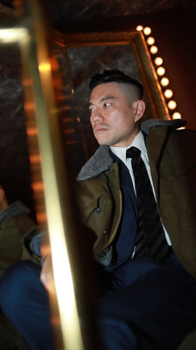 坐在燈泡鏡前的男士,深藍西裝、白襯衫加深色條紋領帶,外面套著毛領軍綠色外套,頭髮兩側推短