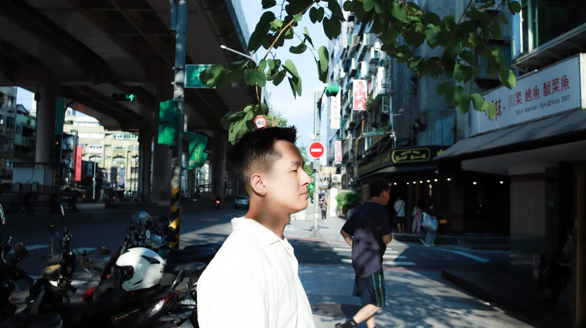 穿白襯衫的男士側臉,站在城市街道的樹蔭下,短髮、側邊收短