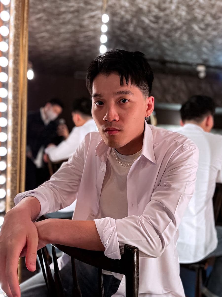 坐在店內燈泡鏡前的男士,白襯衫敞開當外套,內搭米白色圓領上衣,袖口捲到手肘下方